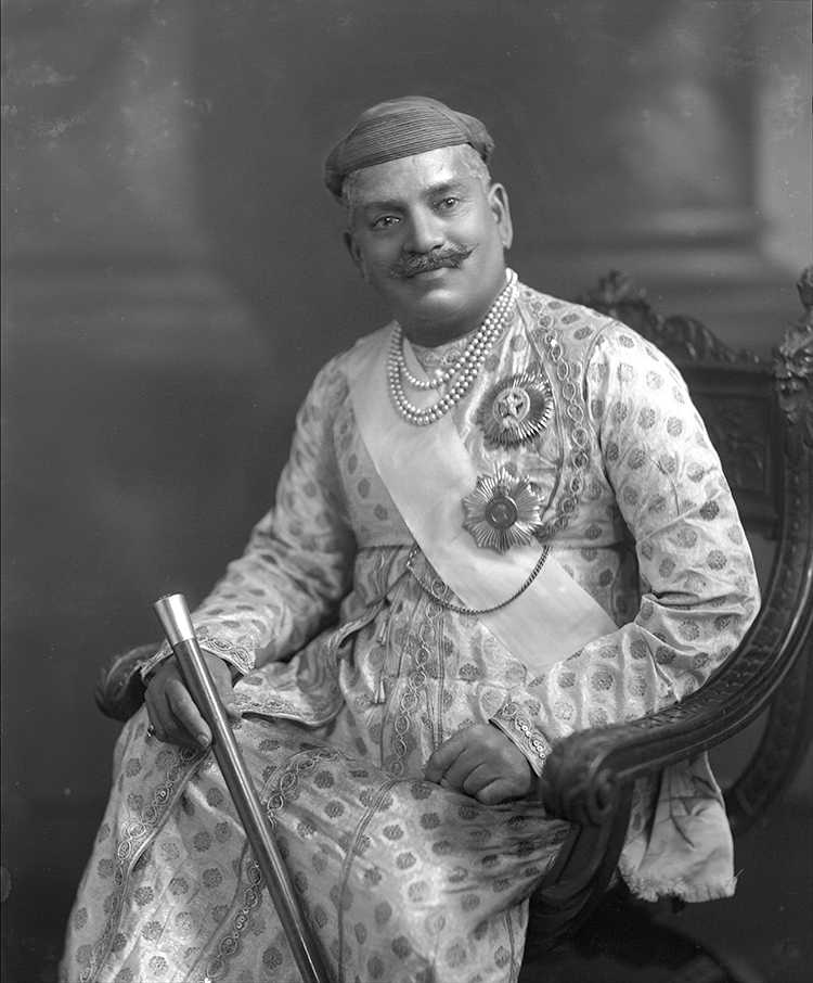 The Gaekwar wears the small, neat turban favoured by the Maratha rulers of western Central India since the early 19th century. This example is unusually plain. He is also wearing a magnificent brocade angarkha (robe). The fabric was probably woven in one of the western Indian centres of textile production such as Ahmedabad or Surat. The design would probably be purple and gold on a yellow or white ground. The robe is embellished with sequins and silk piping around the cuffs and front opening. The angarkha is a traditional man's robe characterised by the round opening on the chest and a somewhat high waist. It is fastened on one side of the neck and tied at the front.